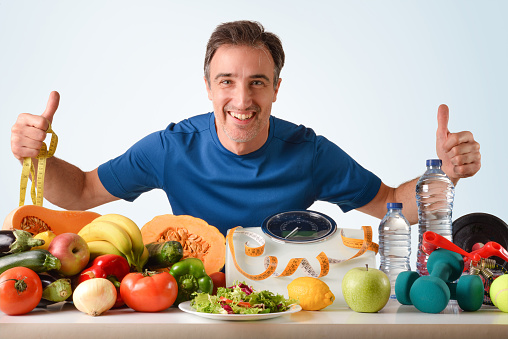 Dietitian athlete presenting healthy food and sport accessories with scale and tape measure for weight control and health with blue isolated background