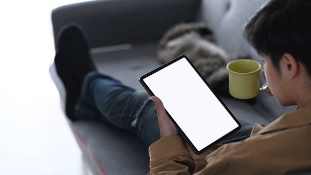 istock Rear view of young man using tablet checking social media and drinking coffee while sitting on sofa at home. 1292904999
