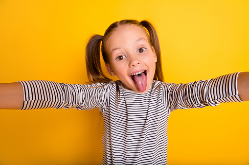 Portrait of young funky fooling playful little girl child kid make selfie stick tongue out isolated on yellow color background.