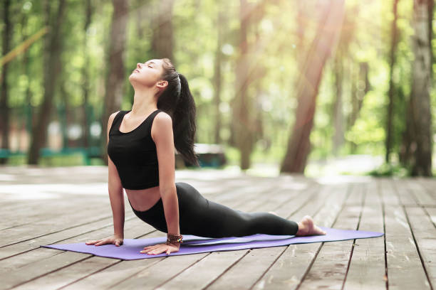 Young brunette woman in sportswear, practicing yoga asanas, performs cobra exercise in the park on a wooden bridge stock photo