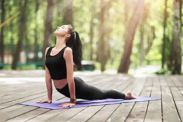 Photo of Young brunette woman in sportswear, practicing yoga asanas, performs cobra exercise in the park on a wooden bridge