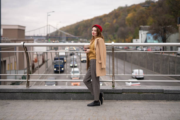 istock Charming french young woman drinks coffee using a coffee mug looking at camera smiling standing at pedestrian bridge with cars on the road. Looking happy fashion girl in a red beret and beige coat 1292912812