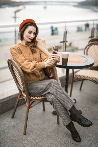 istock Charming french young woman using her smartphone taking selfie while having cup of coffee. Frenchwoman wearing red beret sitting on terrace of restaurant or cafe with background of autumn urban city 1292913595