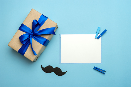 Father's day composition. Gift box with blue ribbon, mustache and blank card on blue background. Top view flat lay with free space for text. Greeting card for dad.