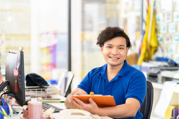 trabalhador de escritório asiático feliz trabalhando na oficina de automóvel, sentado enquanto trabalhava com desktop de computador e prancheta e vestindo uniforme azul. conceito de serviço de carro - car workshop service expertise - fotografias e filmes do acervo