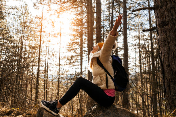 istock Hiking young woman with backpack sitting on rock at woods with hands raised 1292928543
