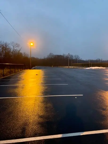 Street light over wet parking lot during rain storm at night with snow pile. Dusk, dark parking spots with forest in background.