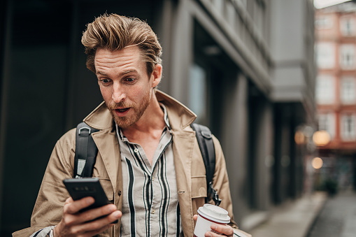 One man, handsome young man on the street outdoors in city, holding disposable cup of coffee and using smart phone.