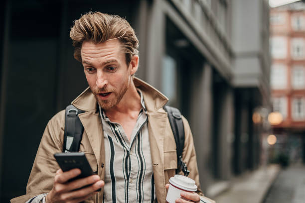 Surprised man using phone on the street in city One man, handsome young man on the street outdoors in city, holding disposable cup of coffee and using smart phone. surprise stock pictures, royalty-free photos & images