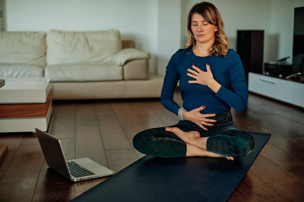 morena de mediana edad sentada en poses de loto y meditando en casa mientras sigue tutoriales en vídeo. - inspirar fotografías e imágenes de stock