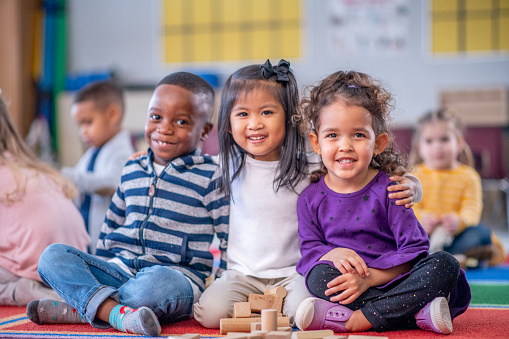 Multi ethnic group of children sitting together in their preschool room. Two girls and a boy smiling widely at the camera while embracing each other. The boy is African American. One girl is mixed raced and the other girl is asian.