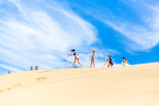 Anna Bay, Australia - December 13, 2020: A teenage girl is about to sandboard down a large sand dune before other tourists. Sandboarding is the most popular activity in Anna Bay.