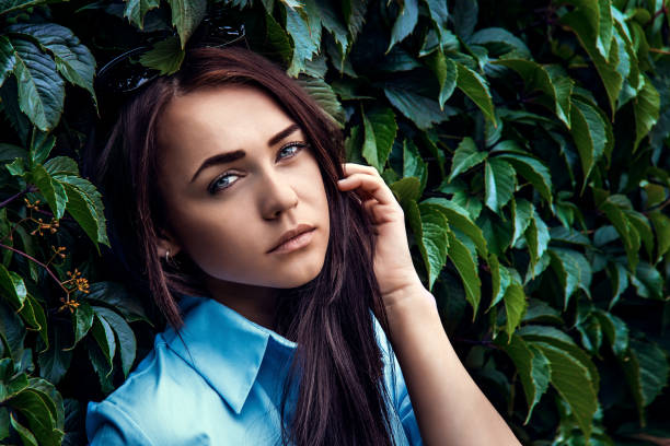 Beautiful Young Girl in Leaves. Handsome Female of European Appearance Wearing Blue Shirt in Green Summer Leaves. Face Beauty Skin. Fashion Shoot.