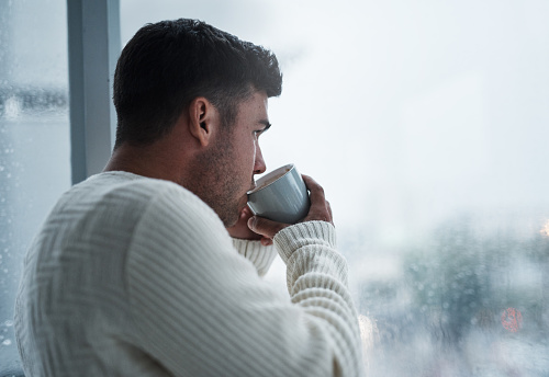 Shot of a young man having coffee and looking out of a window on a rainy day at home