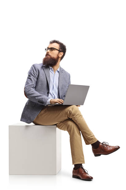 istock Bearded man sitting on a white cube with a laptop computer and looking behind 1293009491