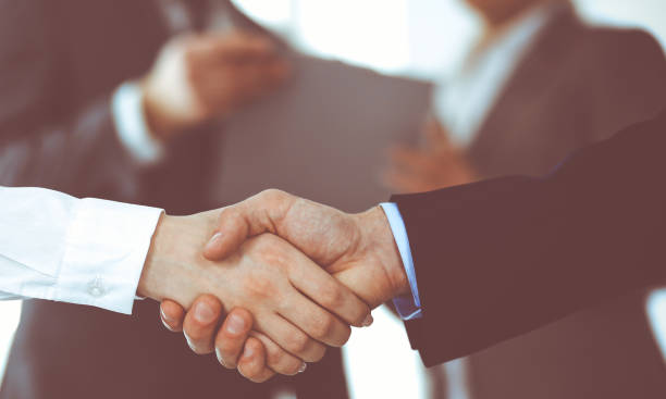 istock Business people shaking hands while standing with colleagues after meeting or negotiation, close-up. Group of unknown businessmen and women in modern office. Teamwork, partnership and handshake concept, toned picture 1293018569