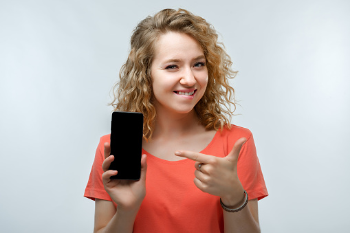 Young woman with curly hairstyle, dressed in casual t-shirt Showing Smartphone Blank Screen, looking at Camera Recommending Mobile App
