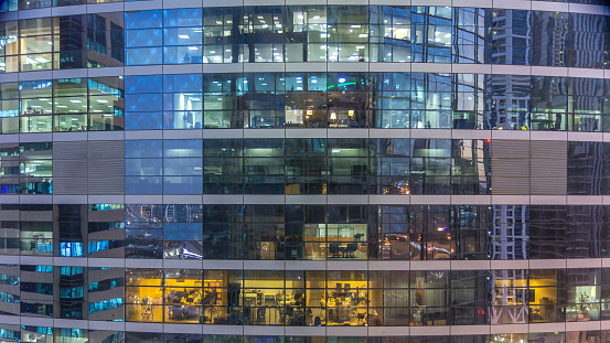 Glowing windows in multistory modern glass and metal office building light up at night timelapse. Workers in a box. Office building illuminated at night. Pan down