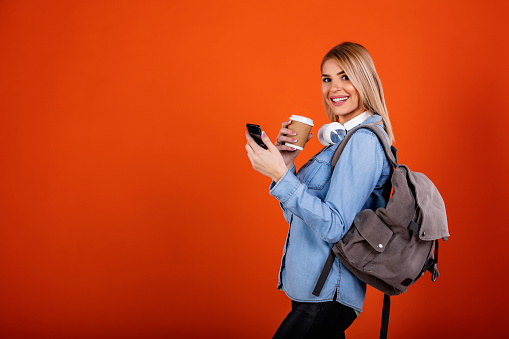 Portrait of Happy young traveler girl holding smartphone and coffee cup, wearing backpack while standing in studio empty red room.