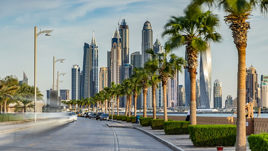 Waterfront promenade on the Palm Jumeirah with palms at road and traffic timelapse before sunset. Dubai, United Arab Emirates. Dubai marina towers on background