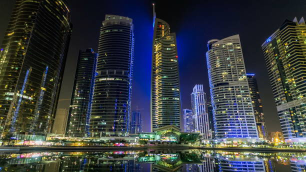 Residential buildings in Jumeirah Lake Towers timelapse hyperlapse in Dubai, UAE Timelapse hyperlapse night view on illuminated skyscrapers at waterfront with palms. Residential buildings in Jumeirah Lake Towers in Dubai, UAE. The JLT is a large development which consists of 79 towers with 3 artificial lakes and park. View from embankment with reflection in water jumeirah stock pictures, royalty-free photos & images
