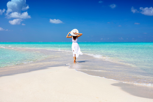 A woman in a white summer dress stands on a sandbar surrounded by turquoise ocean in the Maldives islands