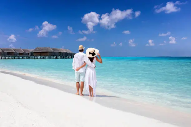 A hugging couple in white summer clothing stands on a tropical beach and enjoys the view to the turquoise sea and blue sky