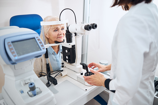 Calm blonde senior woman sitting at the slit lamp with her chin atop the support