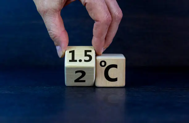Symbol for limiting global warming. Male hand turnes a cube and changes the expression '2 C' to '1.5 C', or vice versa. Concept. Beautiful dak wooden table, black background, copy space.