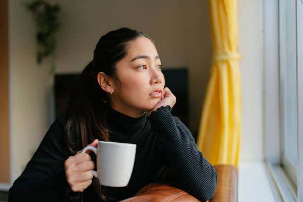 joven mujer sentada en el sofá por ventana y disfrutando de la bebida caliente en casa en la sala de estar - tea women beauty pampering fotografías e imágenes de stock