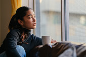 Young woman sitting on sofa by window and enjoying hot drink at home in living room