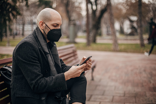 One man, modern young man sitting on bench in park outdoors, he is using mobile phone and wearing protective face mask.