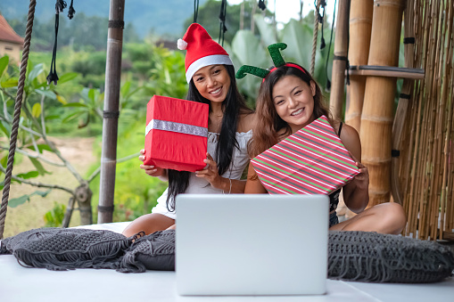Close up shot of two Asian friends showing christmas gifts in front a laptop during christmas web conference at holiday villa