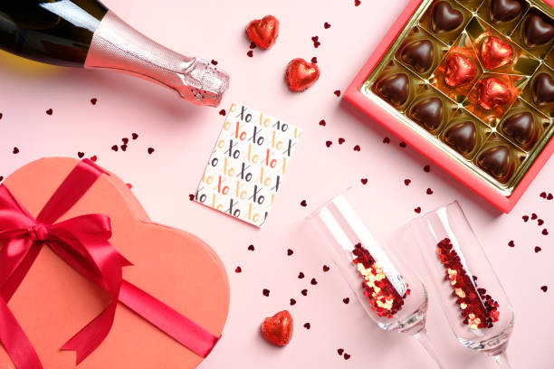 istock Valentines Day flat lay composition. Top view box of chocolates, heart shaped gift box, champagne bottle and glasses, greeting card and confetti on pink table. 1293123866