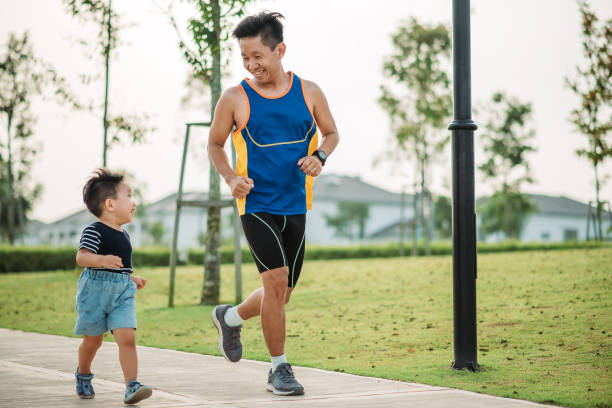Asian chinese smiling father and his son running togethers in the public park during weekend morning stock photo