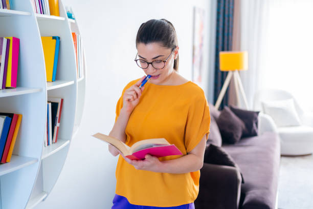 istock Young beautiful woman is reading a book and wearing reading glasses while standing by bookshelf in her living room at home 1293133074