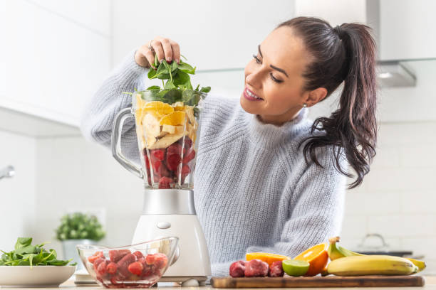 chica de buen aspecto pilas colorida mezcla de frutas y hojas verdes en una licuadora en casa en la cocina. - blender fotografías e imágenes de stock