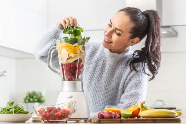 Good looking girl piles colorful mix of fruit and green leaves into a blender at home in the kitchen.