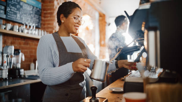 belle barista latino-américaine avec cheveux courts et lunettes fait une tasse de cappuccino savoureux dans coffee shop bar. portrait de happy employee derrière cozy loft-style cafe counter. - bar à expresso photos et images de collection