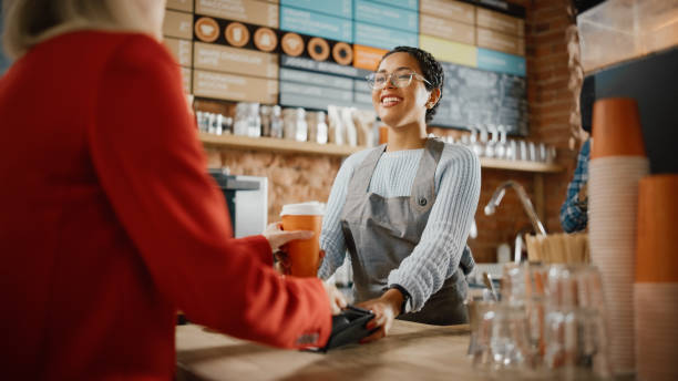 istock Joyful Multiethnic Diverse Woman Gives a Payment Terminal to Customer Using NFC Technology on Smartphone. Customer Uses Mobile to Pay for Take Away Latte and Croissant to a Barista in Coffee Shop. 1293174944