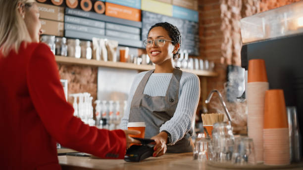 istock Joyful Multiethnic Diverse Woman Gives a Payment Terminal to Customer Using NFC Technology on Smartphone. Customer Uses Mobile to Pay for Take Away Latte and Pastry to a Barista in Coffee Shop. 1293174961