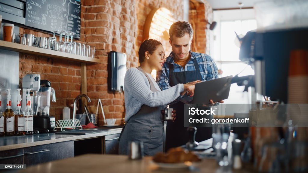 Two Diverse Entrepreneurs Have a Team Meeting in Their Stylish Coffee Shop. Barista and Cafe Owner Discuss Work Schedule and Menu on Laptop Computer. Multiethnic Female and Male Restaurant Employees. Business Stock Photo