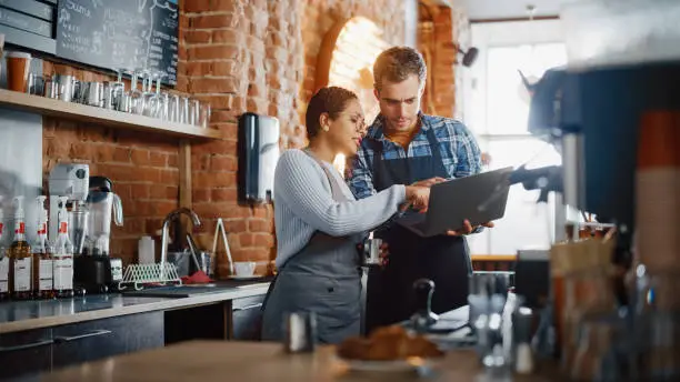 Photo of Two Diverse Entrepreneurs Have a Team Meeting in Their Stylish Coffee Shop. Barista and Cafe Owner Discuss Work Schedule and Menu on Laptop Computer. Multiethnic Female and Male Restaurant Employees.