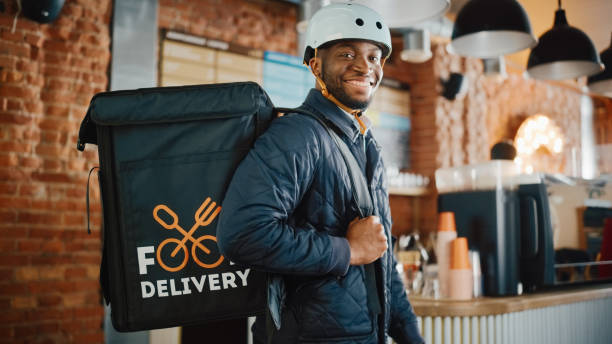 schöne schwarz afroamerikanische lebensmittel lieferung courier posiert vor der kamera in einem coffee shop. glücklicher und lächelnder mann, der einen fahrradhelm und eine wärmegedämmte tasche für essen auf dem rücken trägt. - speisen isoliert stock-fotos und bilder