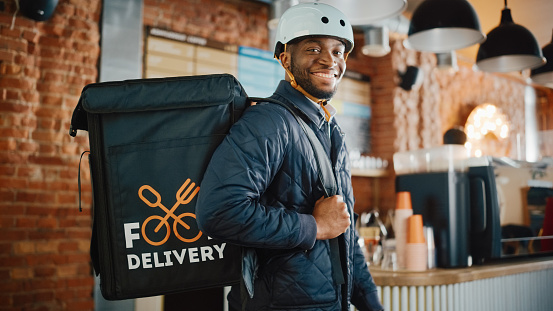 istock Guapo Negro Afroamericano Entrega de Alimentos Courier Posando en frente de la Cámara en una cafetería. Hombre feliz y sonriente con un casco de bicicleta y bolsa con aislamiento térmico para la comida en su espalda. 1293175039