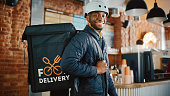 Handsome Black African American Food Delivery Courier Posing in Front of the Camera in a Coffee Shop. Happy and Smiling Man Wearing a Bicycle Helmet and Thermal Insulated Bag for Food on His Back.