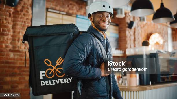 Handsome Black African American Food Delivery Courier Posing In Front Of The Camera In A Coffee Shop Happy And Smiling Man Wearing A Bicycle Helmet And Thermal Insulated Bag For Food On His Back Stock Photo - Download Image Now