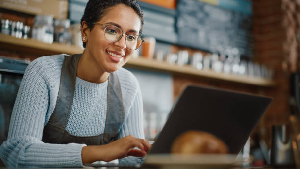 joven y hermosa latina cafetera propietario está trabajando en computadora portátil y comprobar el inventario en un acogedor café. happy restaurant manager o employee browsing internet and chatting with friends. - delicatessen small business cafeteria bar counter fotografías e imágenes de stock