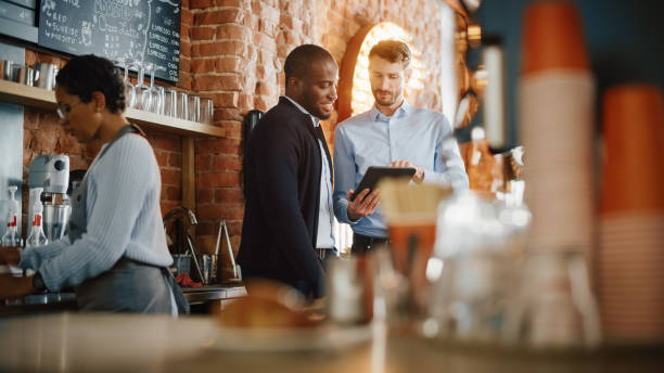 istock Multicultural Coffee Shop Owners Meeting Behind the Counter and Working on Tablet Computer and Checking Inventory in a Cozy Loft-Style Cafe. Successful Restaurant Managers and Barista at Work. 1293175087