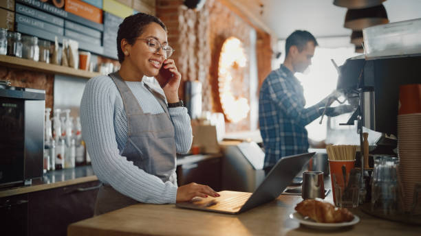 il dipendente della caffetteria latinoamericana accetta un pre-ordine su una telefonata mobile e lo scrive sul computer portatile in un cozy cafe. restaurant manager navigando in internet e parlando su smartphone. - piccola impresa foto e immagini stock